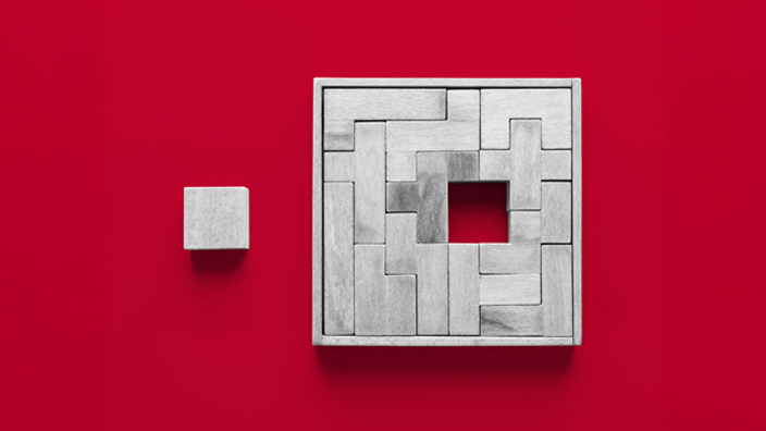 A minimalist image showing a small gray cube placed to the left of a larger square frame filled with interlocking gray rectangular blocks. The larger frame has a square hole in its center, and both objects sit on a bright red background, creating strong contrast.