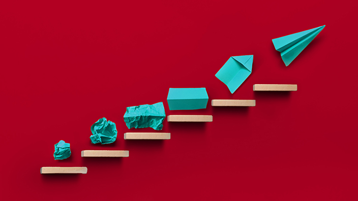 A progression of teal paper shapes arranged on ascending wooden steps against a red background, evolving from crumpled paper to a paper airplane.