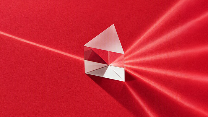 Clear glass prism on a red surface refracting a single beam of light into multiple light beams.