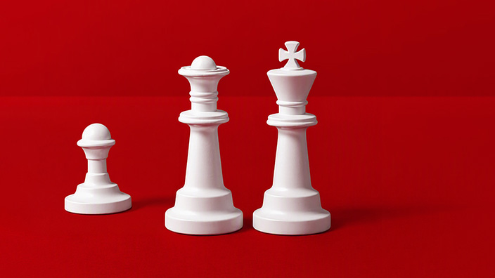 Three white chess pieces stand against a bright red background: a pawn on the left, a queen in the center, and a king on the right. The pieces are arranged in ascending height, with the king as the tallest piece, creating a simple, high-contrast composition.
